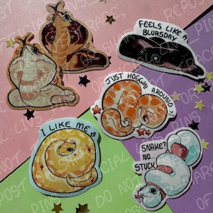 May include: A selection of snake stickers in various colours and designs. One sticker reads "I LIKE ME", another says "FEELS LIKE A BLURSDAY", and another says "JUST HOGGIN AROUND". The stickers are decorated with stars and placed on a pink, green, and purple background.