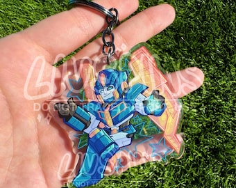 Pode incluir: Um porta-chaves de acrílico transparente com um robô estilizado em azul e amarelo numa pose dinâmica. O design inclui um grande raio laranja atrás do robô e pequenos contornos de estrelas azuis espalhados pela superfície transparente.