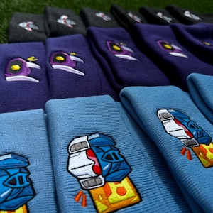 Puede incluir: Una pila de gorros de punto azul y morado con diseños bordados. Los gorros azules presentan un puño de robot con un fondo amarillo y naranja. Los gorros morados presentan una cabeza de robot morado y blanco con un fondo amarillo.