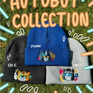 May include: A collection of three beanies with embroidered cartoon robot designs. The beanies are in blue, gray, and black. The blue beanie says "PRIME" and the black beanie says "BEE". The text "AUTOBOT COLLECTION" is at the top.