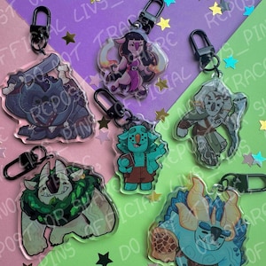 Puede incluir: Una colección de llaveros acrílicos con ilustraciones de personajes de fantasía coloridos. Cada llavero tiene un cierre negro y representa una criatura o figura única con detalles vibrantes. El fondo incluye confeti en forma de estrella y texto.