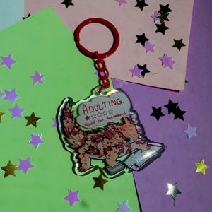 May include: A clear acrylic keychain with a red ring and chain. The keychain features a cartoon lizard with the text "ADULTING. Would Not Recommend". The lizard is brown, orange, and white. The background is green and purple with star-shaped confetti.