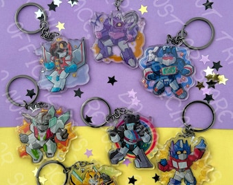 May include: Seven acrylic keychains featuring stylized robot characters in various poses and colors, including red, blue, yellow, purple, and green. Each keychain has a black metal ring and is surrounded by small star-shaped confetti pieces.