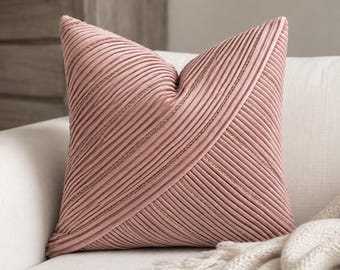 Puede incluir: Un cojín cuadrado de color rosa empolvado con un diseño de tela plisada y texturizada. La superficie está cubierta por filas diagonales de pliegues de tela suaves y elevados, con finas costuras en tono dorado metálico entre los pliegues.
