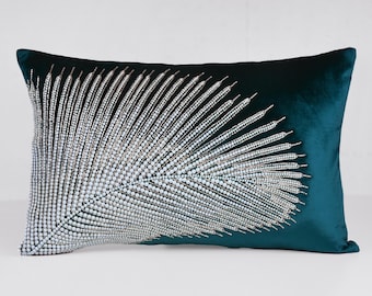 May include: Teal velvet rectangular pillow with a beaded, stylized feather design. The feather is made of small, iridescent beads that catch the light. The pillow is a decorative accent for a bed or sofa.