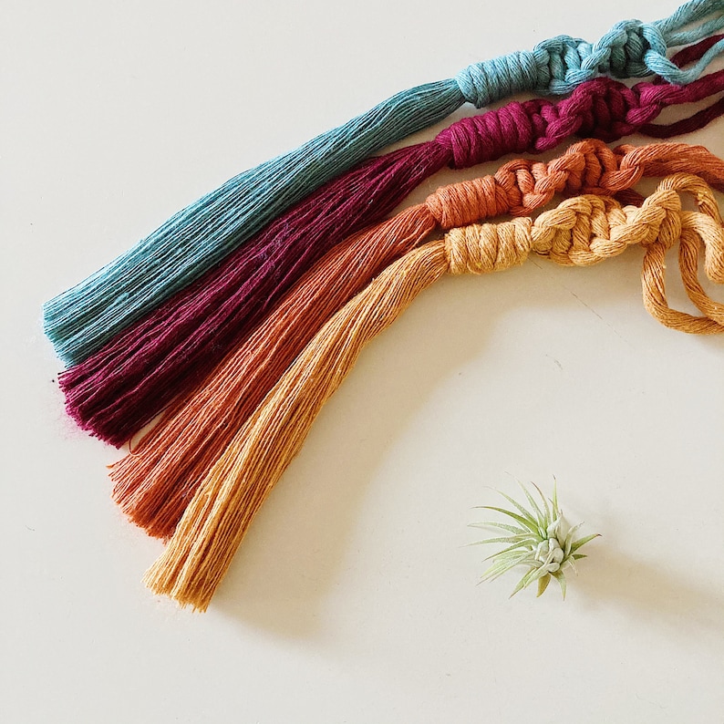 May include: Four colorful macrame tassels in shades of teal, burgundy, orange, and yellow. The tassels are arranged in a rainbow pattern and are tied together at the top.