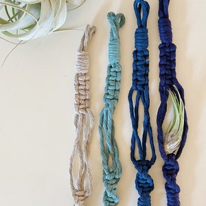 May include: Four macrame plant hangers in different shades of blue and beige. The hangers have a knotted design and a tassel at the bottom.