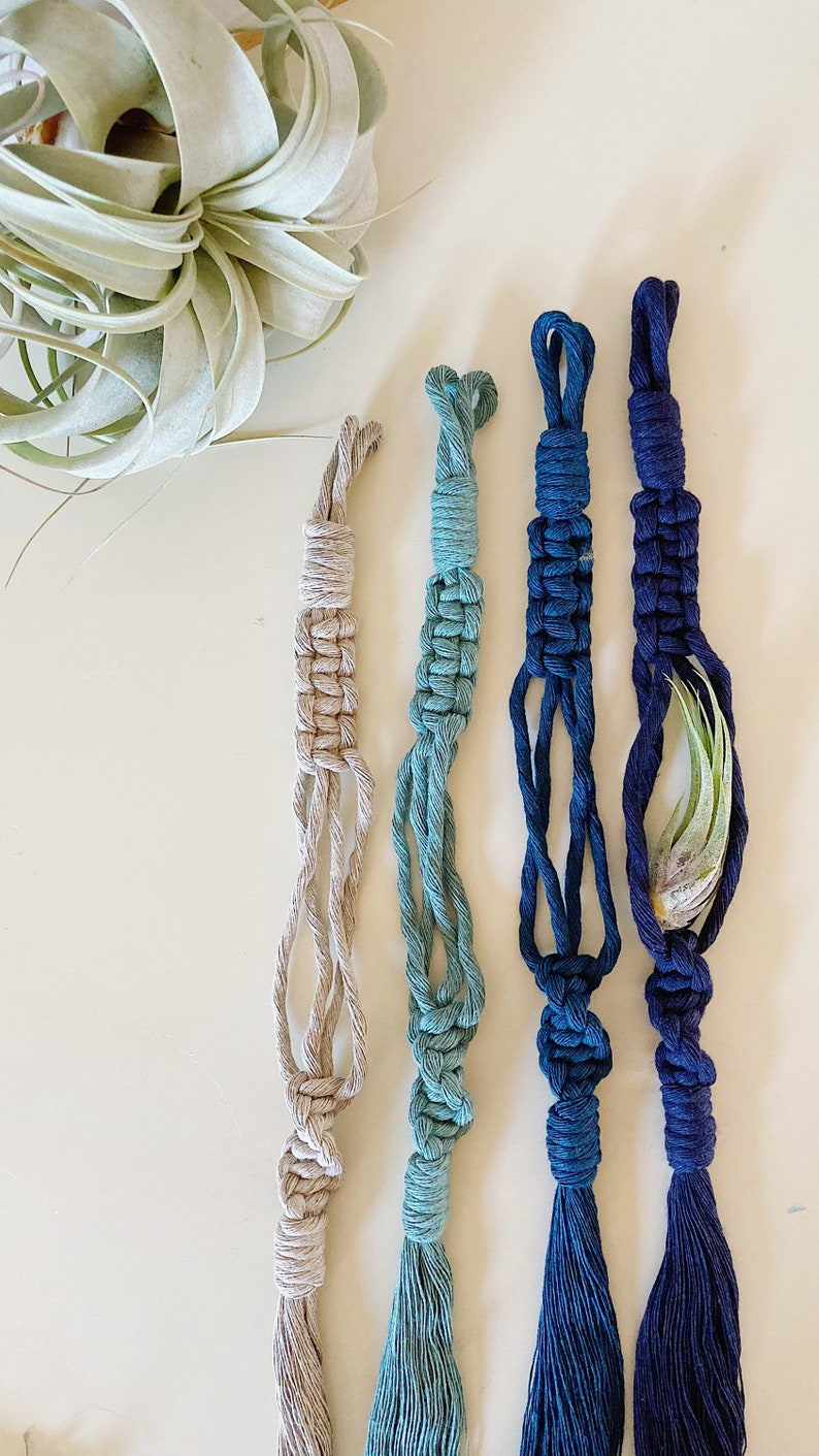 May include: Four macrame plant hangers in different shades of blue and beige. The hangers have a knotted design and a tassel at the bottom.