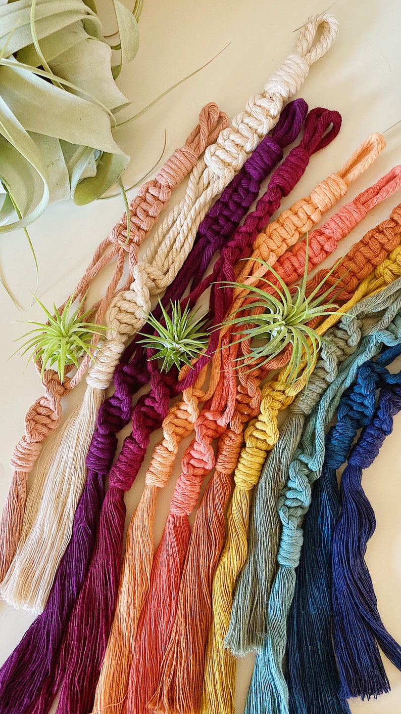 May include: A collection of colorful macrame plant hangers with tassels. The hangers are made with natural cotton cord and feature a variety of colors, including natural, burgundy, orange, yellow, green, and blue.
