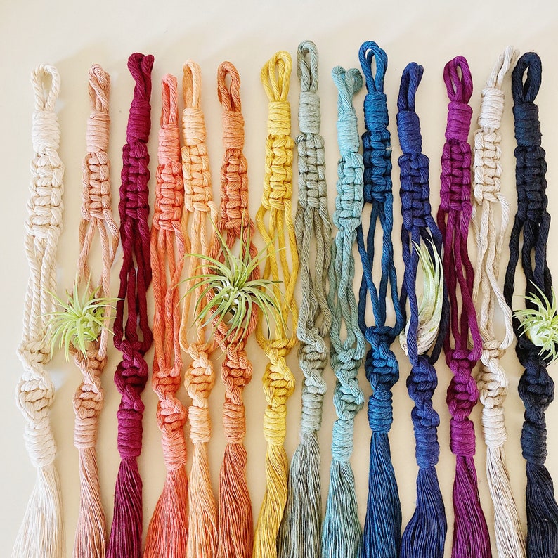 May include: A set of 14 macrame plant hangers in various colors, including white, beige, red, orange, yellow, green, blue, and purple. Each hanger features a knotted design and a tassel at the bottom.