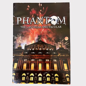 May include: A red and black poster for the Phantom of the Opera Las Vegas Spectacular. The poster features a dramatic image of the Phantom's mask and a view of the Paris Opera House.