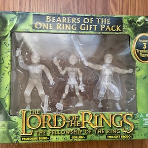 May include: A box set of three translucent action figures from The Lord of the Rings: The Fellowship of the Ring. The figures are Frodo, Gollum, and Bilbo. The box features a green background with the movie title and a warning label.