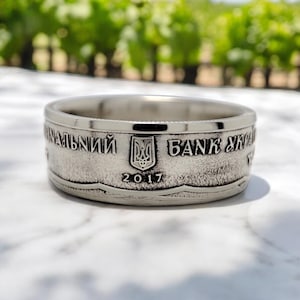 Pode incluir: Um anel de prata feito com uma moeda ucraniana. O anel apresenta o brasão de armas ucraniano, o texto "Національний банк України" (Banco Nacional da Ucrânia) e o ano "2017".
