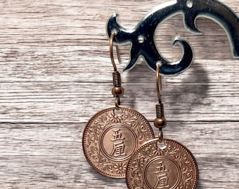 May include: Copper coin earrings with a floral design and Asian characters. The earrings hang from copper-colored ear wires and are displayed against a wooden background.