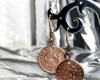 May include: Copper coin earrings with intricate designs and a vintage aesthetic. The earrings hang from copper-colored hooks and feature detailed engravings on the circular coins. The earrings are displayed against a shiny, silver background.