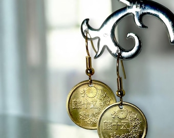 May include: A pair of gold-toned coin earrings. Each earring features a circular coin with Japanese characters and floral detailing. The earrings hang from gold-colored hooks and are displayed against a blurred background.