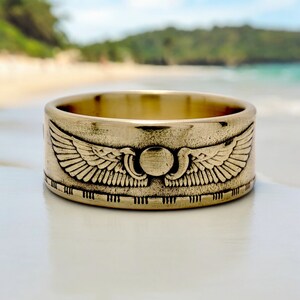 May include: A gold-colored ring with a raised design of two wings and a circle in the center. The ring is on a white surface.