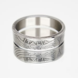 Op de afbeelding: Een zilveren ring gemaakt van twee gestapelde Italiaanse Lira-munten. De munten tonen de woorden "ITALIA" en een gevleugeld ontwerp.