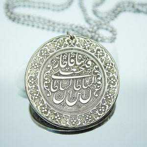 May include: A silver pendant necklace with a round, ornate silver charm featuring intricate Arabic calligraphy. The charm is surrounded by a decorative border.
