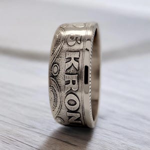 Op de afbeelding: Een zilveren ring gemaakt van een 5 Kronor-munt. De munt heeft een ontwerp van concentrische cirkels en de tekst "5 KRONOR".