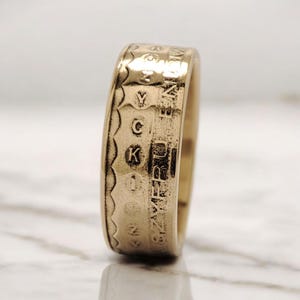 May include: A gold-colored ring with engraved letters and numbers. The ring has a textured surface with a series of engraved letters and numbers around the band. The ring is displayed on a marble surface.