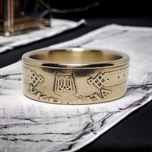 Pode incluir: Um anel dourado com um design em relevo que apresenta o brasão de armas da Ucrânia, a palavra "UKRAINE" em escrita cirílica e um padrão tradicional ucraniano.