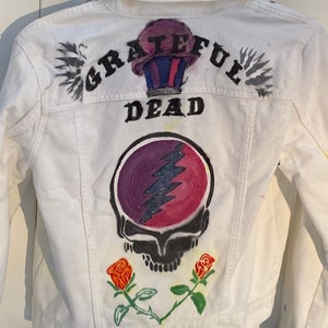 Peut inclure: Veste en jean blanche avec un motif Grateful Dead peint à la main. Le motif comprend les mots "Grateful Dead" et un crâne avec un éclair. Il y a aussi deux roses peintes en bas.