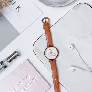 May include: A brown leather wristwatch with a white face and a butterfly design. The watch has a gold-tone case and hands.