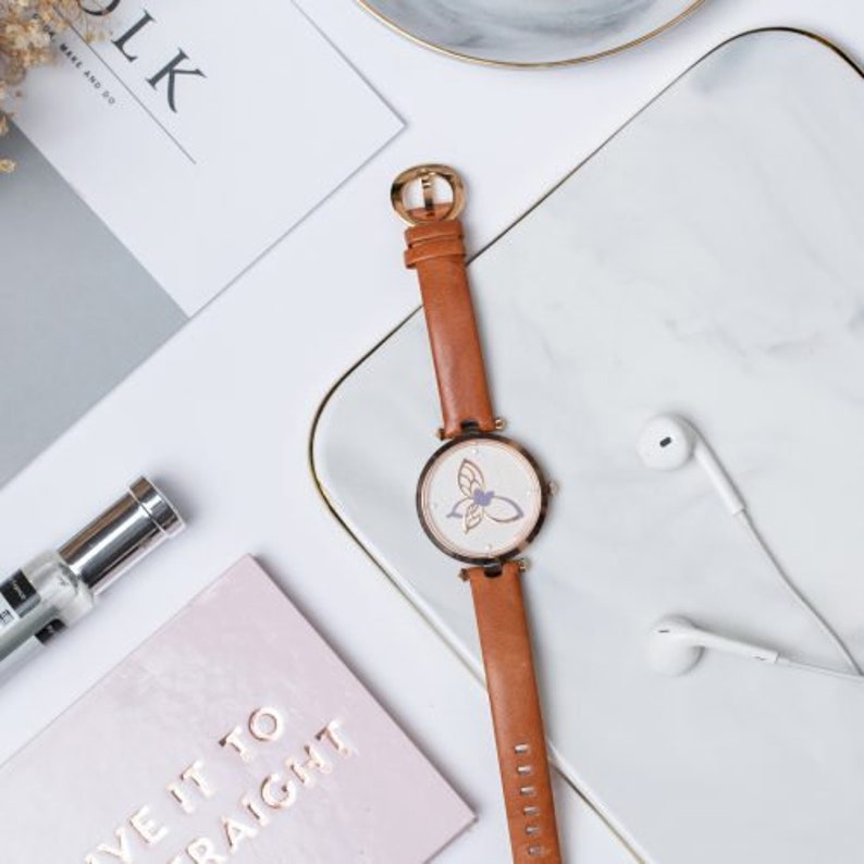 May include: A brown leather wristwatch with a white face and a butterfly design. The watch has a gold-tone case and hands.