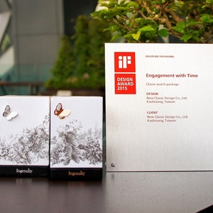 May include: Two white boxes with a black and white floral design and a silver and gold butterfly on each box. The boxes are labeled "Ingenuity". The boxes are sitting on a wooden table in front of a silver award plaque with the text "iF DESIGN AWARD 2015" and "Engagement with Time".