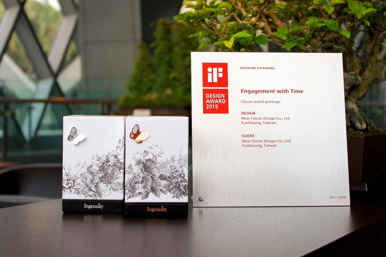 May include: Two white boxes with a black and white floral design and a silver and gold butterfly on each box. The boxes are labeled "Ingenuity". The boxes are sitting on a wooden table in front of a silver award plaque with the text "iF DESIGN AWARD 2015" and "Engagement with Time".