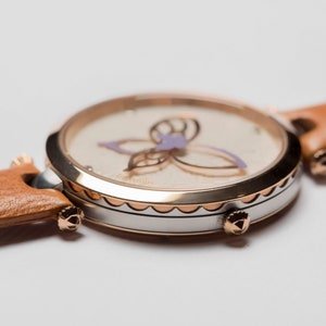 May include: A round, silver and rose gold watch with a white face and a brown leather band. The face features a delicate butterfly design with purple accents.