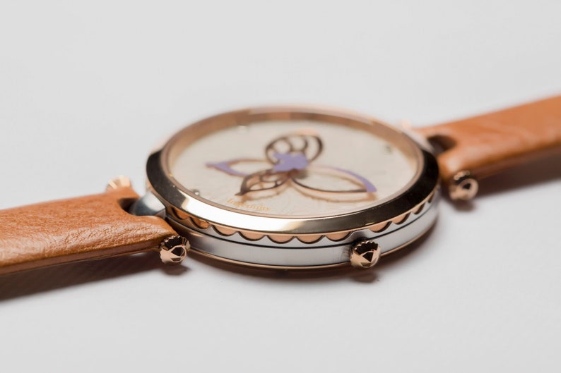 May include: A round, silver and rose gold watch with a white face and a brown leather band. The face features a delicate butterfly design with purple accents.