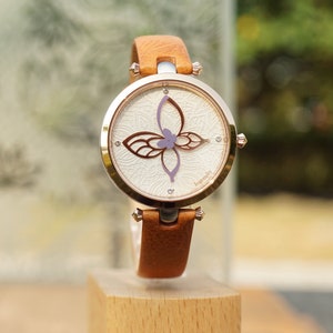 May include: A brown leather wristwatch with a white face and a delicate butterfly design. The watch face has a white background with a delicate floral pattern and the word "Ingenuity" printed on the bottom right corner.