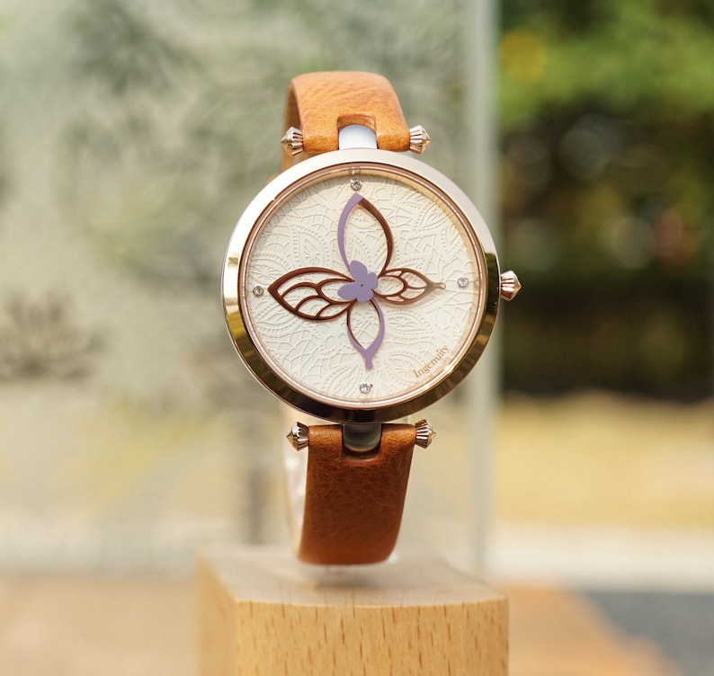 May include: A brown leather wristwatch with a white face and a delicate butterfly design. The watch face has a white background with a delicate floral pattern and the word "Ingenuity" printed on the bottom right corner.
