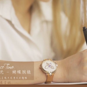 May include: A gold-tone wristwatch with a white face and a butterfly design. The watch has a brown leather band. The text "Elegance Time" is visible in the image.