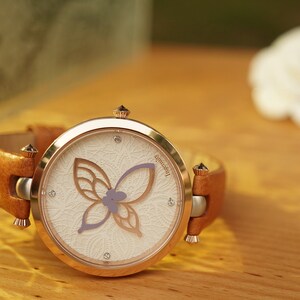 May include: A rose gold wristwatch with a white face and a brown leather band. The watch face features a delicate butterfly design with a purple flower.