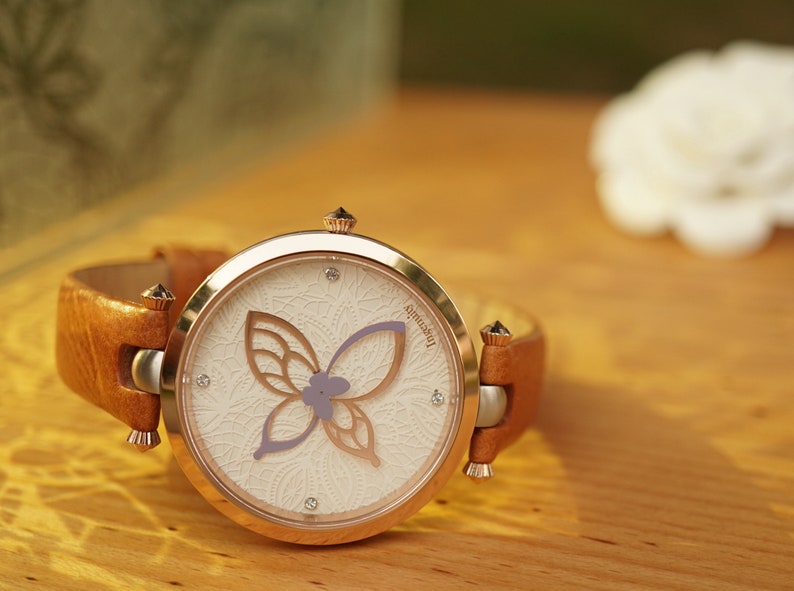 May include: A rose gold wristwatch with a white face and a brown leather band. The watch face features a delicate butterfly design with a purple flower.