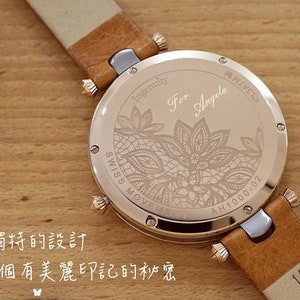 May include: A gold-toned wristwatch with a brown leather band. The back of the watch has an engraved design of a floral pattern and the text "Ingenuity For Angela AH1000-02".