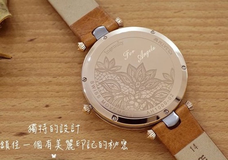 May include: A gold-toned wristwatch with a brown leather band. The back of the watch has an engraved design of a floral pattern and the text "Ingenuity For Angela AH1000-02".