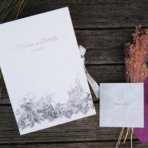 May include: A white card with the text "Memories on Butterfly" and "Ingenuity" printed in a cursive font. The card is decorated with a black and white floral design. A small white card with the text "Ingenuity" is also visible. A brown leather watch with a butterfly design is on a purple cloth.