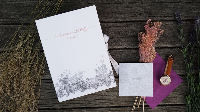 May include: A white card with the text "Memories on Butterfly" and "Ingenuity" printed in a cursive font. The card is decorated with a black and white floral design. A small white card with the text "Ingenuity" is also visible. A brown leather watch with a butterfly design is on a purple cloth.