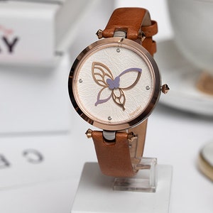 May include: A brown leather wristwatch with a rose gold case and a white face featuring a purple butterfly design. The watch face also has a small logo that says "Ingenuity".