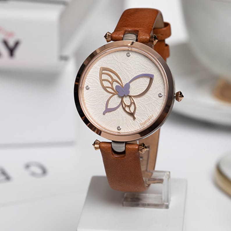 May include: A brown leather wristwatch with a rose gold case and a white face featuring a purple butterfly design. The watch face also has a small logo that says "Ingenuity".