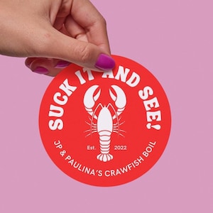 May include: A red circular sticker with the text "Suck It And See!" in white. The sticker also features a white crawfish illustration and the text "JP & Paulina's Crawfish Boil Est. 2022".