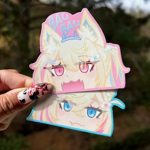 May include: Two anime-style stickers featuring a blonde character with pink and blue accents. The top sticker has pink eyes and the text "BAU BAU". The bottom sticker has blue eyes. The stickers are held up by a hand with a black and red nail design.