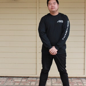 May include: A person wearing a black long-sleeved shirt with white text that reads "YOUR VIBE ATTRACTS YOUR TRIBE" on the right sleeve and black pants. The person is wearing gray sneakers.