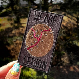 May include: A black and gray embroidered patch with the text "We Are Legion" and a brown, stitched, circular design with a red line.