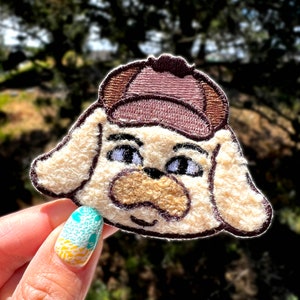 May include: A white and brown embroidered patch of a dog wearing a brown hat. The dog has a brown nose and brown eyes. The patch is made of a soft, fuzzy material.
