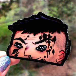 May include: A sticker of a cartoon character with black hair and a bloody face. The character has a dark expression and is looking to the right. The sticker is white with black outlines and has the text "wtfmr-shop" in the bottom right corner.